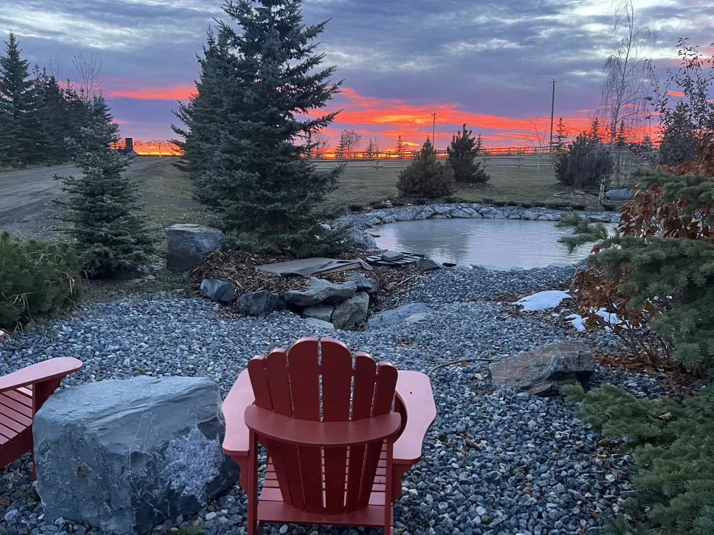 Pond with two Adirondack chairs and orange sunset.
